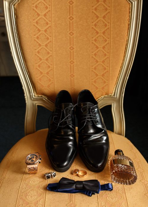 Wedding groom accessories on chair: black shoes, wrist watch, perfume bottle, bow tie near wedding rings